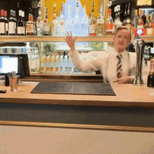 a bartender in a white shirt and tie stands behind a counter with bottles of martini on it