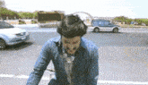 a man in a denim shirt is walking down a street with a car in the background .