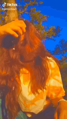 a girl in a tie dye shirt is sitting in front of a tree and taking a selfie .