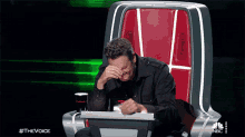 a man is sitting in a chair with his hand on his face and the words #thevoice on the bottom right