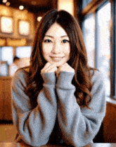 a woman wearing a grey sweater is smiling with her hands on her chin
