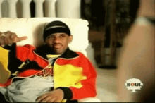 a man in a colorful jacket is sitting on a couch and giving a thumbs up .