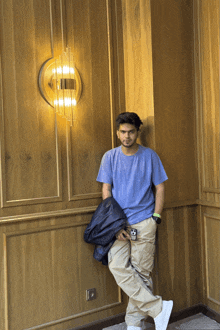 a man wearing a blue shirt and khaki pants leans against a wooden wall