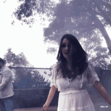 a woman in a white dress is dancing in front of trees and a man in a white suit