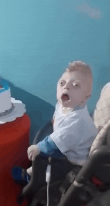 a baby is sitting in a stroller with his mouth open and a cake in the background .