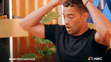 a man in a black shirt holds his hands to his head with the nbc destination x logo behind him