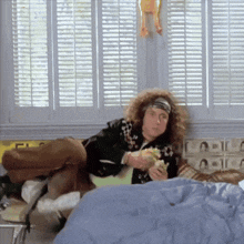 a man with curly hair is sitting on a bed eating a sandwich and playing a guitar