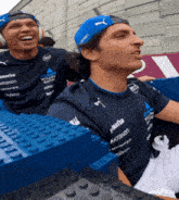 a man wearing a blue hat that says kraken on it sits next to another man