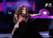 a woman singing into a microphone while holding a purple stuffed animal with the letters ae on the bottom right