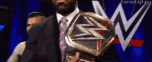 a man in a suit and tie is holding a wrestling championship belt in front of a wwe logo .