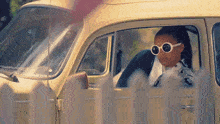 a woman wearing sunglasses is sitting in a yellow car behind a picket fence .