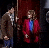 a man in a brown suit and a woman in a red jacket are standing in front of a door