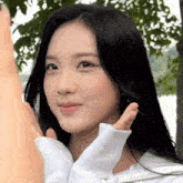 a woman with long black hair is wearing a white sweater and making a heart shape with her hands .
