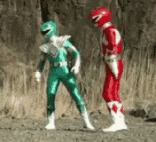 a green power ranger and a red power ranger are standing next to each other on a dirt field .