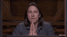 a man with long hair and black nails is sitting at a table with his hands folded .
