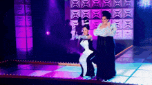 two drag queens are dancing on a stage with purple lights behind them