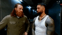 two men are standing next to each other in a room and talking . one of the men is wearing a white tank top .
