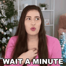 a woman in a pink sweater says wait a minute in front of a christmas tree