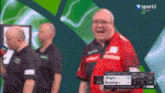 a man in a red shirt is laughing in front of a screen that says v sport2 show