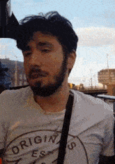 a man with a beard wearing a white shirt that says origins