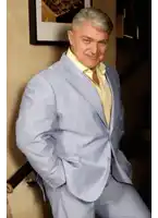 a man in a light blue suit and yellow shirt is leaning against a wall