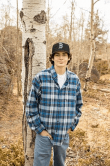 a young man wearing a plaid shirt and a hat with the letter b on it is standing next to a tree .