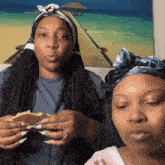 two women wearing headbands are eating sandwiches in front of a painting of a beach