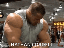 a man with a lot of muscles is sitting in a gym with the name nathan cordell written on the bottom .