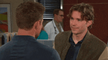 a man in a bomber jacket talks to another man in a blue shirt