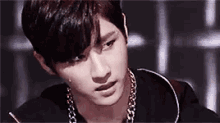 a young man wearing a black shirt and a chain around his neck is looking at the camera .