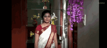 a woman in a white and red saree is standing in front of a shelf with purple flowers ..