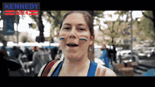 a woman with blue and red stripes painted on her face is smiling in front of a sign that says kennedy 2024