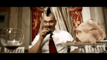 a man with a mohawk is drinking a glass of wine while sitting at a table with a ham .