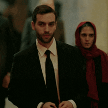 a man in a suit and tie with a woman in a red hijab behind him