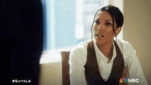 a woman in a white shirt and brown vest is sitting in front of a window with a nbc logo in the corner