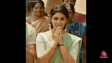 a woman in a green and white saree is standing in front of a crowd of people with her hands folded in prayer .