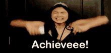 a woman wearing a tiara is celebrating with her arms in the air and the words `` achieveee '' written on the screen .