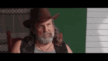 a man with long hair and a beard is wearing a cowboy hat and sitting in a chair .