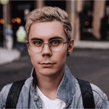 a young man wearing glasses and a denim jacket is looking at the camera