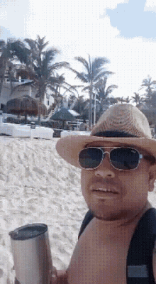 a man wearing a straw hat and sunglasses is holding a tumbler on the beach