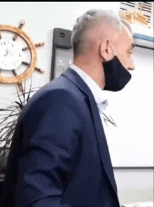 a man in a suit wearing a black face mask stands in front of a clock