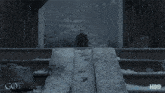 a woman in a black dress is standing in front of a stone wall in a snowy scene from game of thrones .