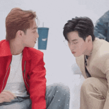 a man in a red jacket and a man in a tan suit are sitting next to each other and looking at each other .