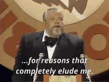 a man in a tuxedo stands at a podium with the words " for reasons that completely elude me " behind him