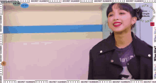 a woman wearing a black jacket and a purple shirt is smiling in front of a sign that says secret number x