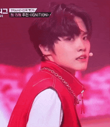 a young man wearing a red vest and a chain around his neck is standing in front of a microphone on a stage .