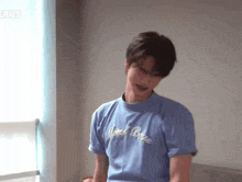 a young man wearing a blue t-shirt is standing in front of a window in a room .