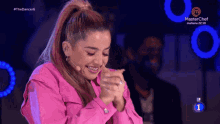 a woman in a pink jacket is smiling and clapping in front of a microphone on a masterchef tv show