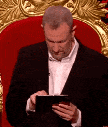 a man in a suit and white shirt is sitting on a red throne looking at his cell phone .