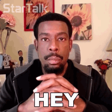 a man with a beard says " hey " with his hands folded in front of him